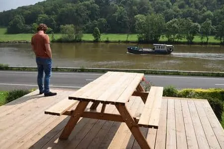 Terrasse en bois avec table de pique-nique face à la Meuse, péniche au passage